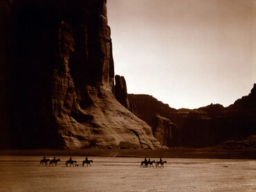 Seven riders on horseback and dog trek against background of canyon cliffs, by Edward Curtis