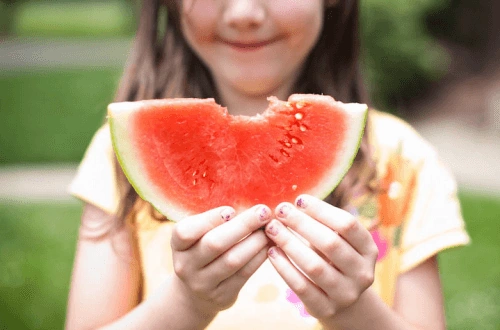 A girl holds a piece of watermelon with a nice bite take out of the edge of it.
