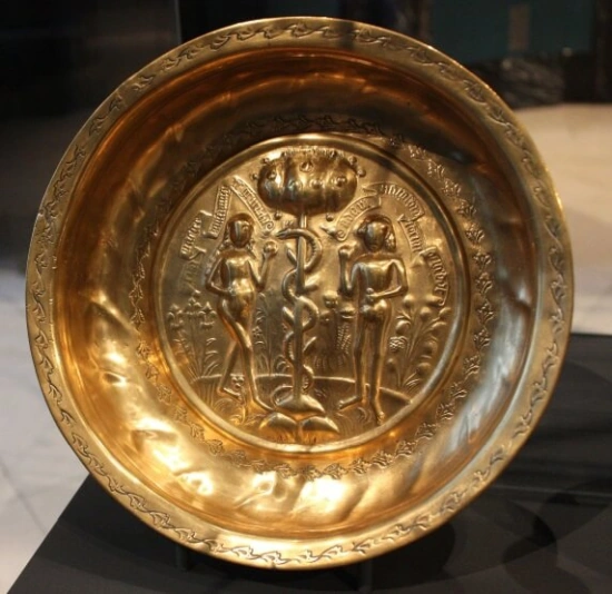 This hand beaten brass bowl, dating from 1500-1550, shows Adam, Even and the Serpent in the Garden of Eden. It is made with repousse and chased brass, in 
Germany - possibly Nuremburg. Both the central design and the decoration upon the rim of this dish were made using a series of stamps impressed into the metal. Nuremberg trade regulations stated that all punches and stamps had to be applied by hand. The scene depicts the Fall of Man, when Adam and Eve were tempted by the serpent to pick an apple from the Tree of Knowledge. Scenes like this one were popular on brass dishes of the 16th century as they added a decorative element to objects for household use. This dish was probably used to wash hands, yet contemporary paintings show that dishes were also displayed upon dressers when not in use.
Collection ID: 454-1907
This photo was taken as part of Britain Loves Wikipedia in February 2010 by Valerie McGlinchey.